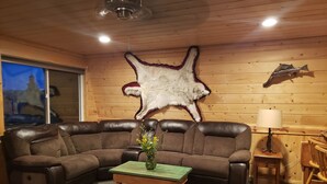 A cozy living room with a brown leather sectional sofa, a wooden coffee table with a vase, a mounted fur rug, a lamp, and a wooden chair. - Charming Lakeview Cabin at Panguitch Lake (Panguitch)