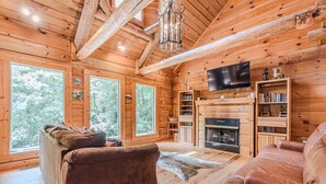 Interior acogedor de una cabaña de madera con chimenea, estanterías de madera y un ventanal con vistas a un bosque.
