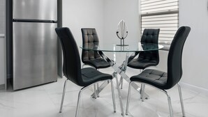 A modern kitchen with a stainless steel refrigerator, a glass dining table with four black chairs, and a pendant light.