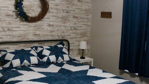 A bedroom with a bed, a nightstand, a wall-mounted light fixture, a curtain, and a decorative wreath. - Clifford's Country Cottage (Bonne Terre)