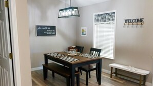 A dining area with a wooden table, chairs, a bench, a hanging light fixture, a 'WELCOME' sign, a framed quote, and a window with blinds. - Clifford's Country Cottage (Bonne Terre)