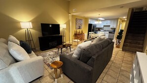 A living room with a sofa, TV, and floor lamp. There is a kitchen in the background.