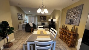 A dining area with a long table, white chairs, and a chandelier. There is a sofa and a television in the background.