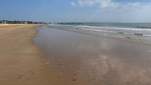 Ein weiter Sandstrand mit sanften Wellen, einem klaren Himmel und Gebäuden in der Ferne entlang der Küste.