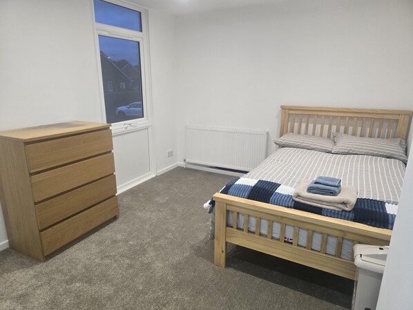 A bedroom with a wooden bed, a wooden dresser, a radiator, and a window with a view of houses.