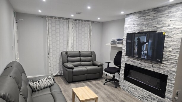 Ett modernt vardagsrum med en grå soffa, en läderfåtölj, ett träbord och en tv som är monterad på en vägg med stenliknande struktur.