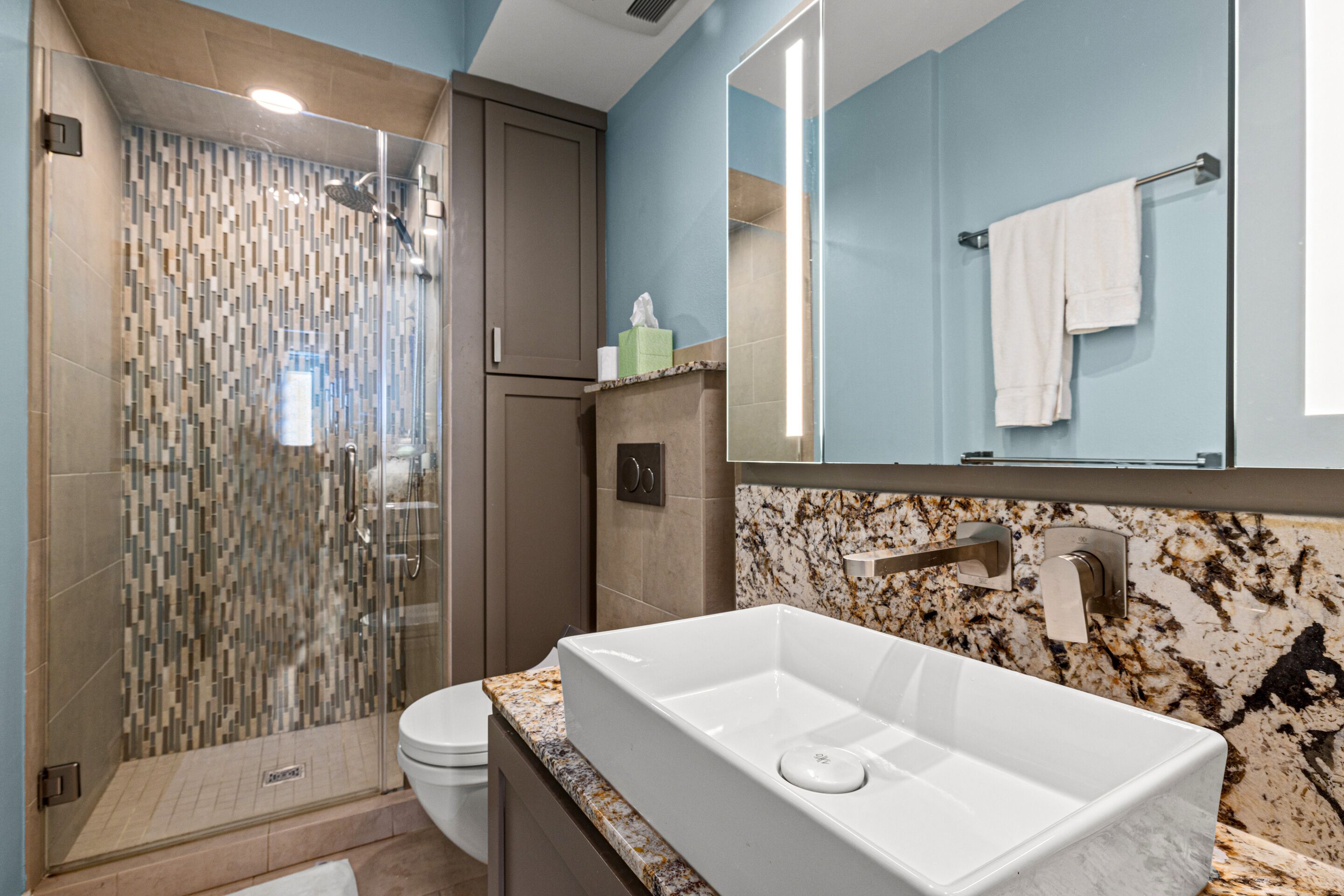 A modern bathroom with a glass-enclosed shower, a white sink with a marble countertop, and a large mirror above the sink.