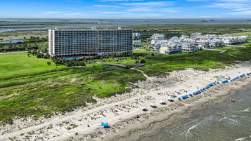 A coastal hotel with a large building, a beach with umbrellas, and a view of the ocean and ships in the distance.