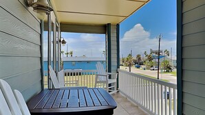 A balcony with a table and chairs, a view of a blue building, and a street with palm trees. - Casa Palmera by Silver Sands Vacation Rentals (Port Aransas)