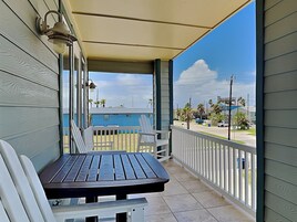 A balcony with a table and chairs, a view of a blue building, and a street with palm trees.