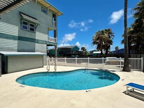 A swimming pool with a white fence, a bench, and a building with a balcony in the background. - Casa Palmera by Silver Sands Vacation Rentals (Port Aransas)