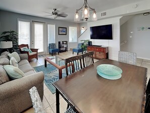 A spacious living room with a wooden dining table, comfortable seating, a ceiling fan, and a large flat-screen TV. - Casa Palmera by Silver Sands Vacation Rentals (Port Aransas)