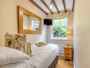 A bedroom with a bed, a bedside table, a lamp, a window with a view of greenery, a mounted TV, and a wooden ceiling beam.