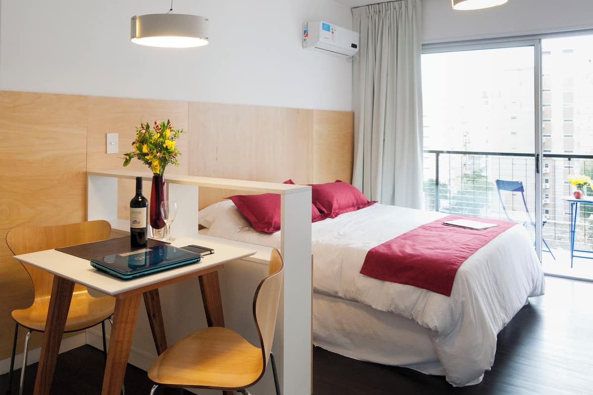 A hotel room with a bed, a desk with a laptop, a chair, a vase with flowers, and a window with a view of buildings.