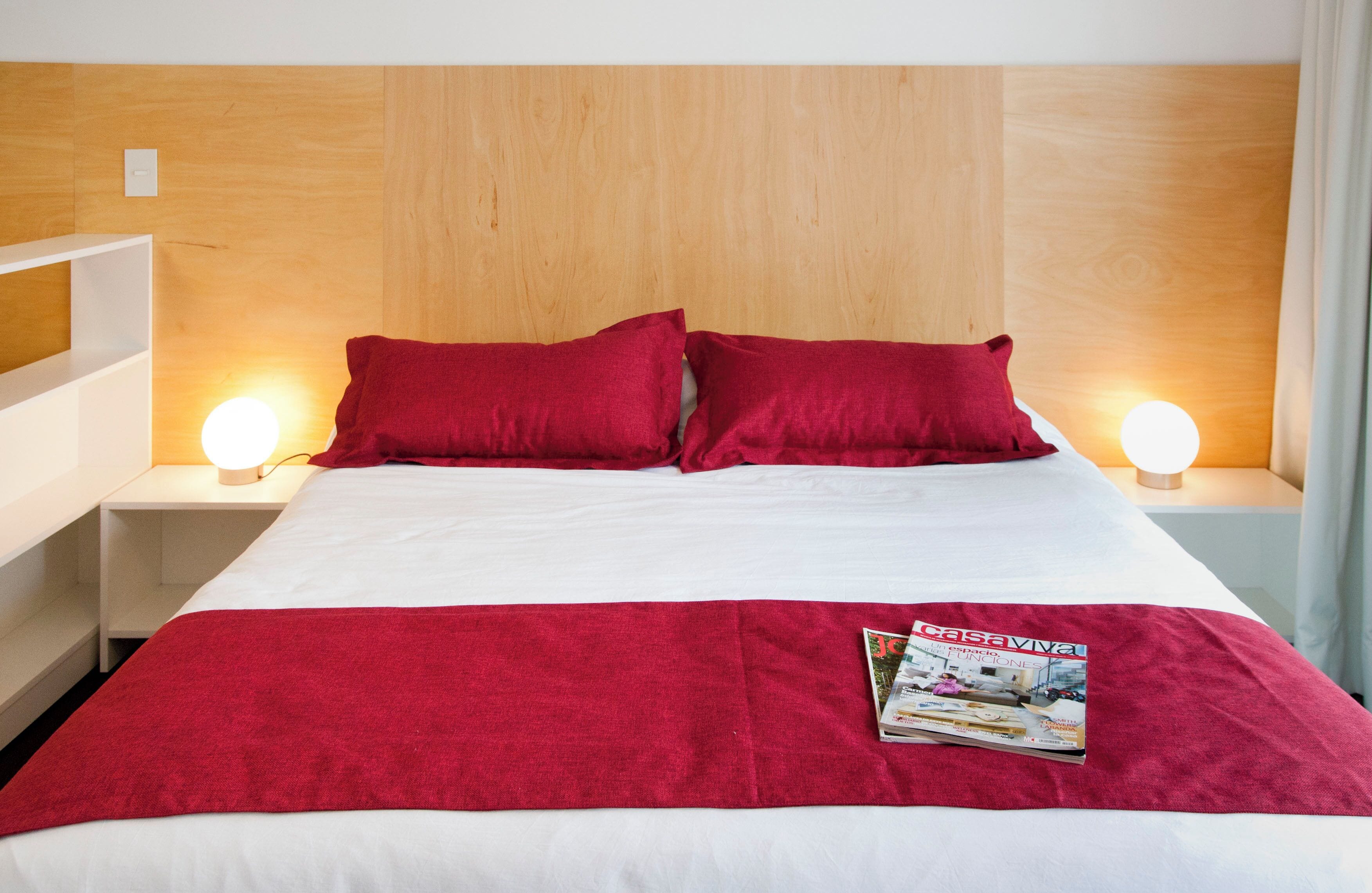 A hotel room with a wooden headboard, white bedding, and red pillows. There is a magazine on the bed and two bedside lamps.