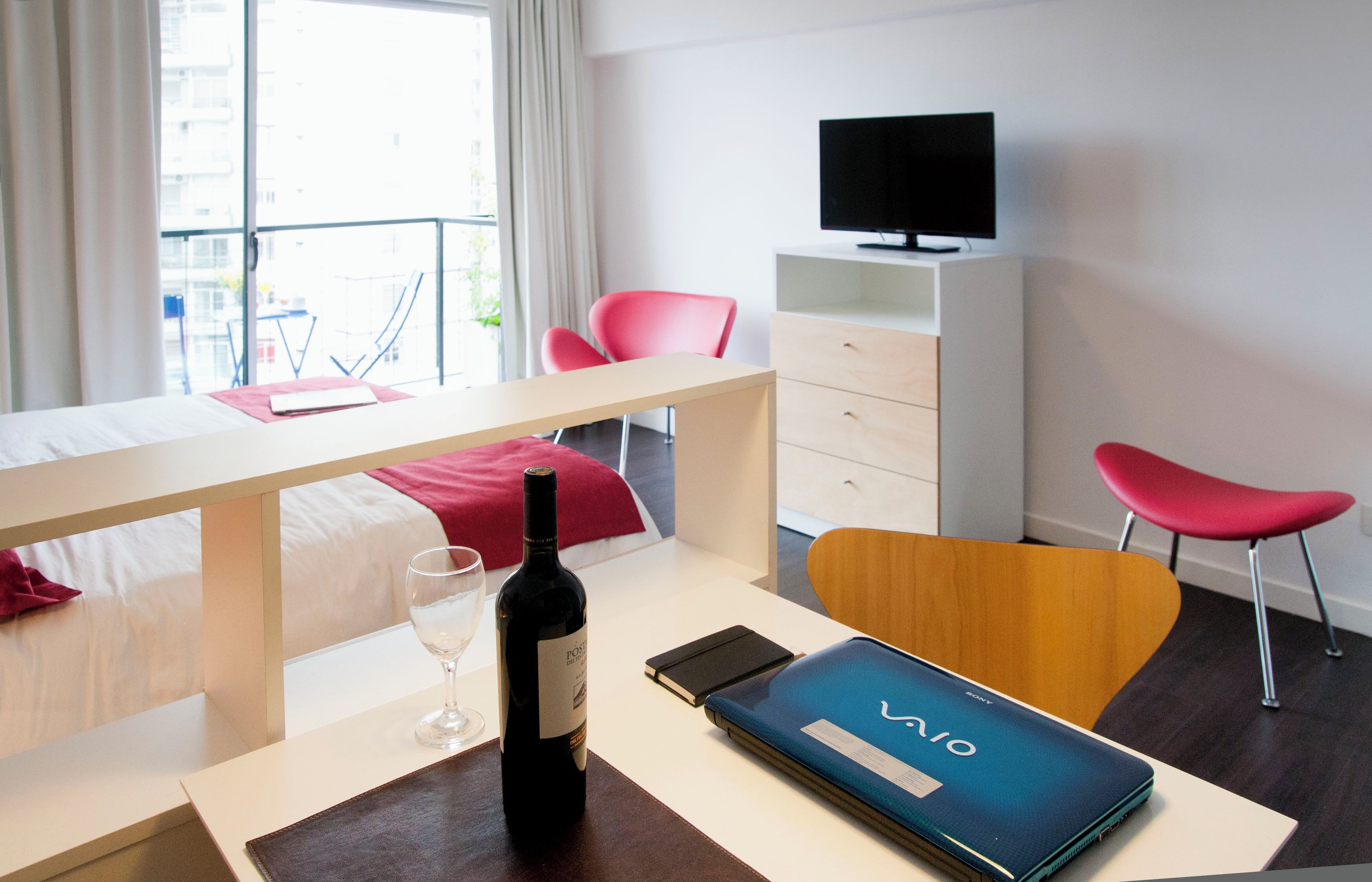 Une chambre d’hôtel avec un lit, un bureau avec un ordinateur portable, une bouteille de vin, une télévision et deux chaises rouges.
