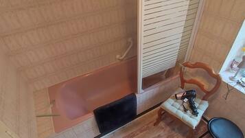 A bathroom with a bathtub, a wooden chair, a small table with a hairdryer, and a window with blinds.