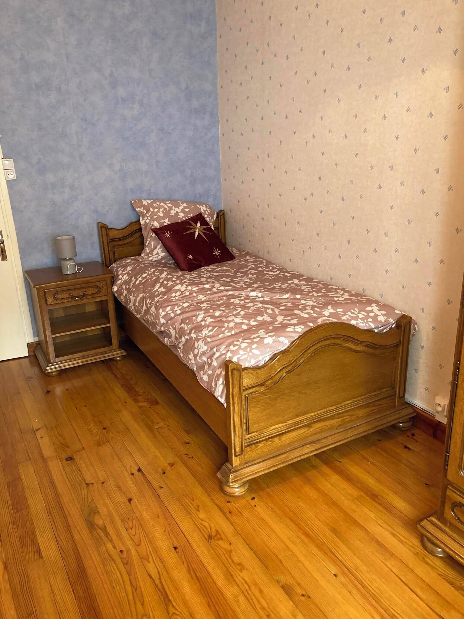 Un lit simple avec un cadre en bois, une table de chevet avec une lampe et un mur décoré d’un motif.