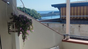 Un balcon avec un canapé et un fauteuil en osier blanc, une jardinière suspendue et une vue sur la mer et les bâtiments en arrière-plan.