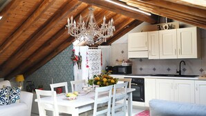 Une cuisine chaleureuse aménagée dans les combles, avec des poutres en bois, un lustre, des meubles blancs et un coin repas avec une table dressée pour deux personnes.