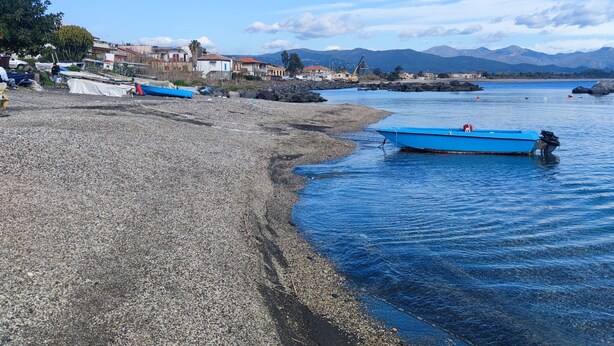 Une plage de galets bordée de bateaux, un ciel dégagé et des montagnes à l’horizon.