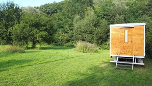 Una estructura de madera con puerta y escalera, ubicada en un campo cubierto de hierba y árboles densos al fondo.