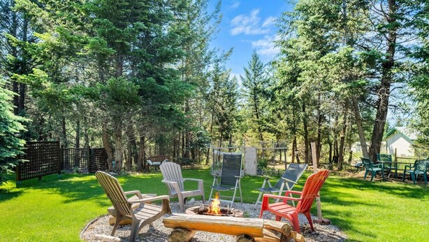 Ein Garten mit Feuerstelle, umgeben von Stühlen und einem Tisch, in einer grünen Rasenfläche mit Bäumen im Hintergrund.