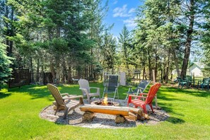 A backyard with a fire pit surrounded by chairs and a table, set in a grassy area with trees in the background. - Wild Embers | Central | Fire Pit | Fireplace (Invermere)