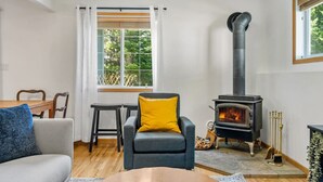 Ein gemütliches Wohnzimmer mit Sofa, Sessel und Tisch. In der Ecke steht ein Kaminofen, und es gibt ein Fenster mit Vorhängen.