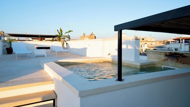 Una terraza en la azotea con piscina, sillas de descanso blancas y una pérgola.