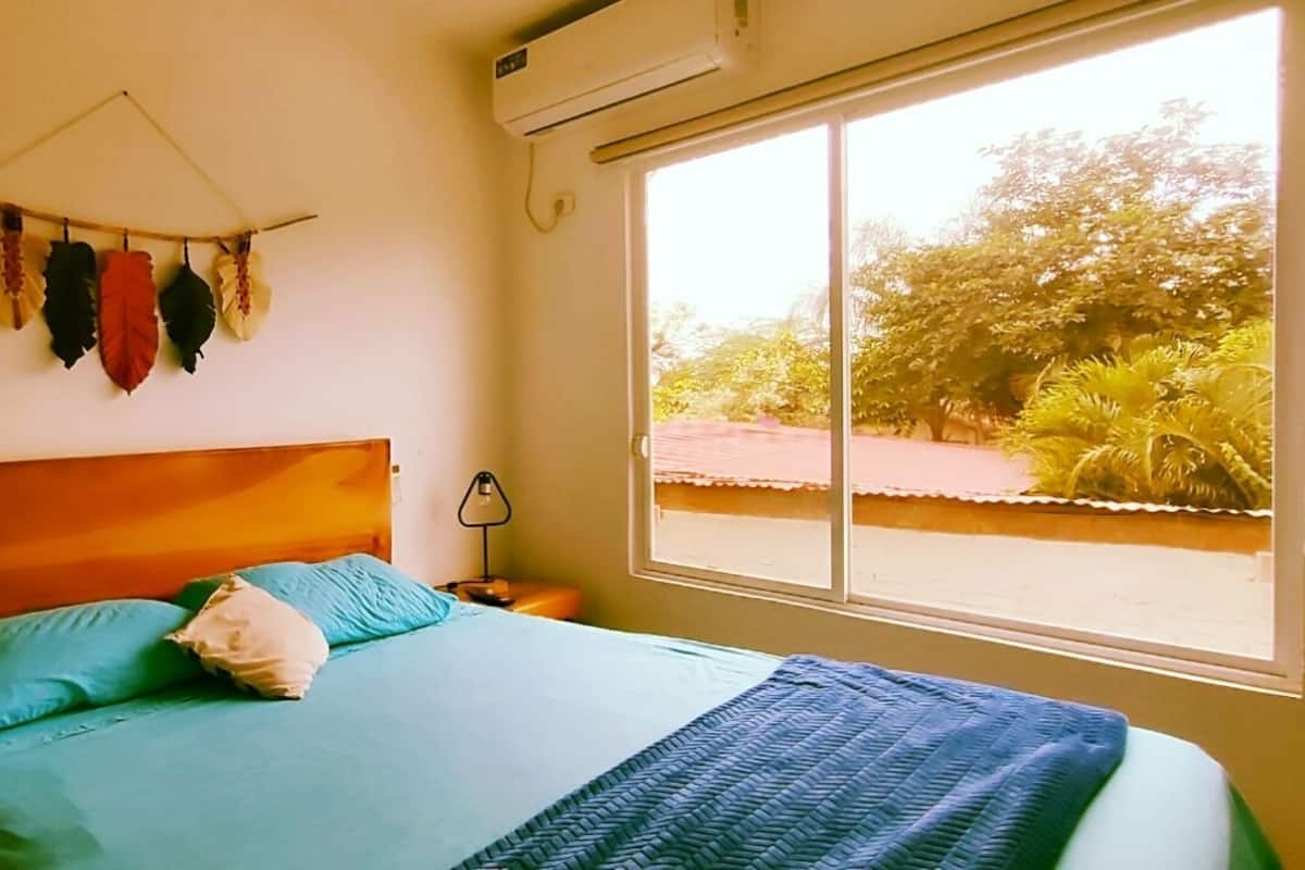 A bedroom with a bed, pillows, a wall-mounted air conditioner, a window with a view of greenery, and a wall decoration.