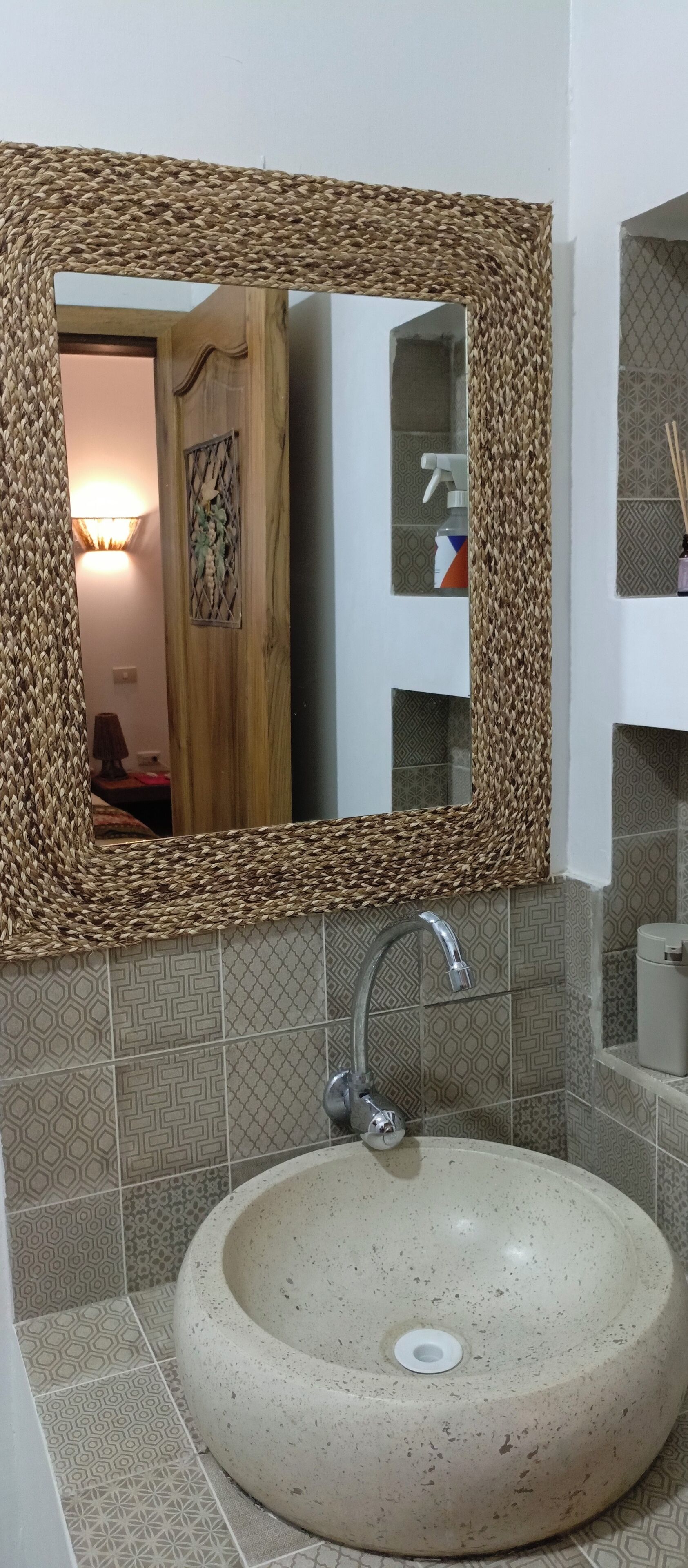 Un baño con un lavabo redondo, un espejo con marco tejido y azulejos estampados.