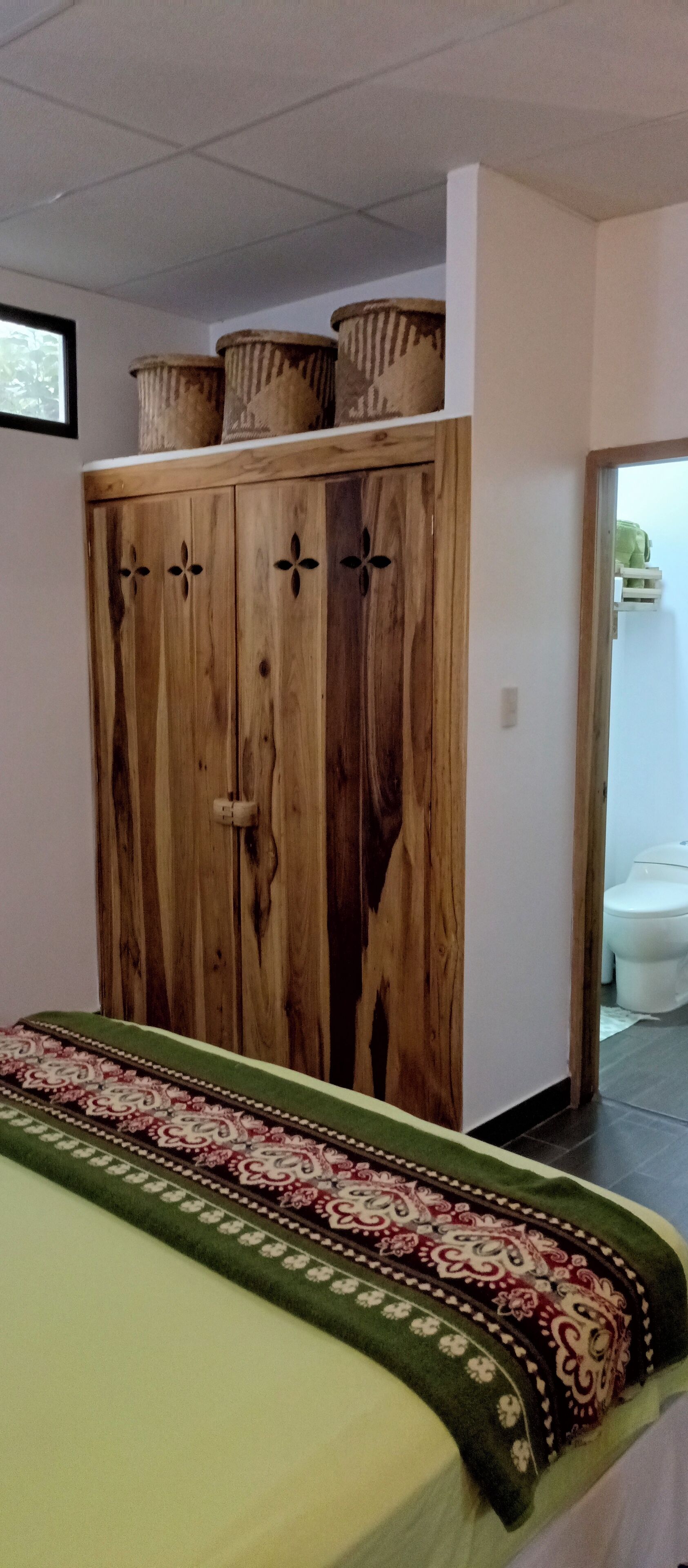 A room with a wooden wardrobe, a bed with a patterned bedspread, and a toilet visible through an open doorway.
