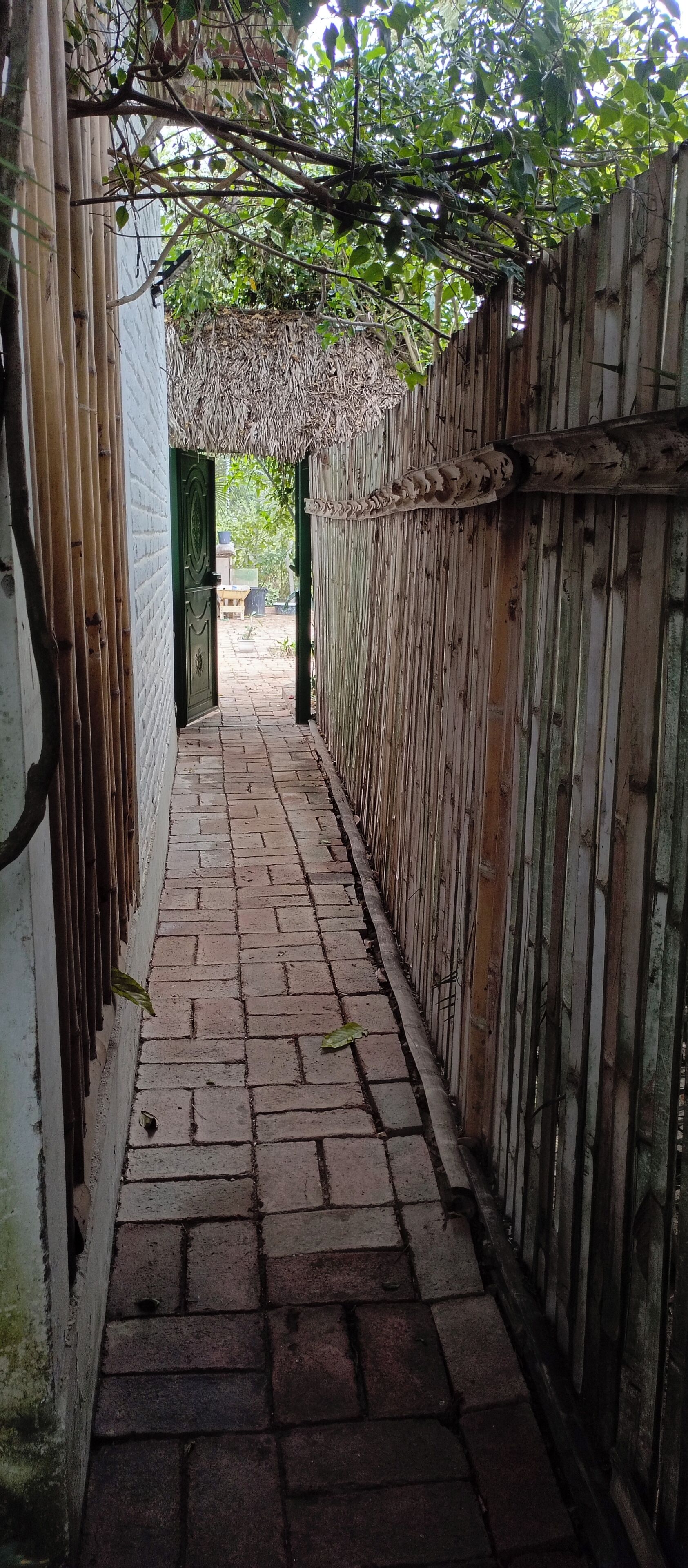 Un étroit sentier pavé de briques, bordé de clôtures en bambou, mène à une construction à toiture de chaume.