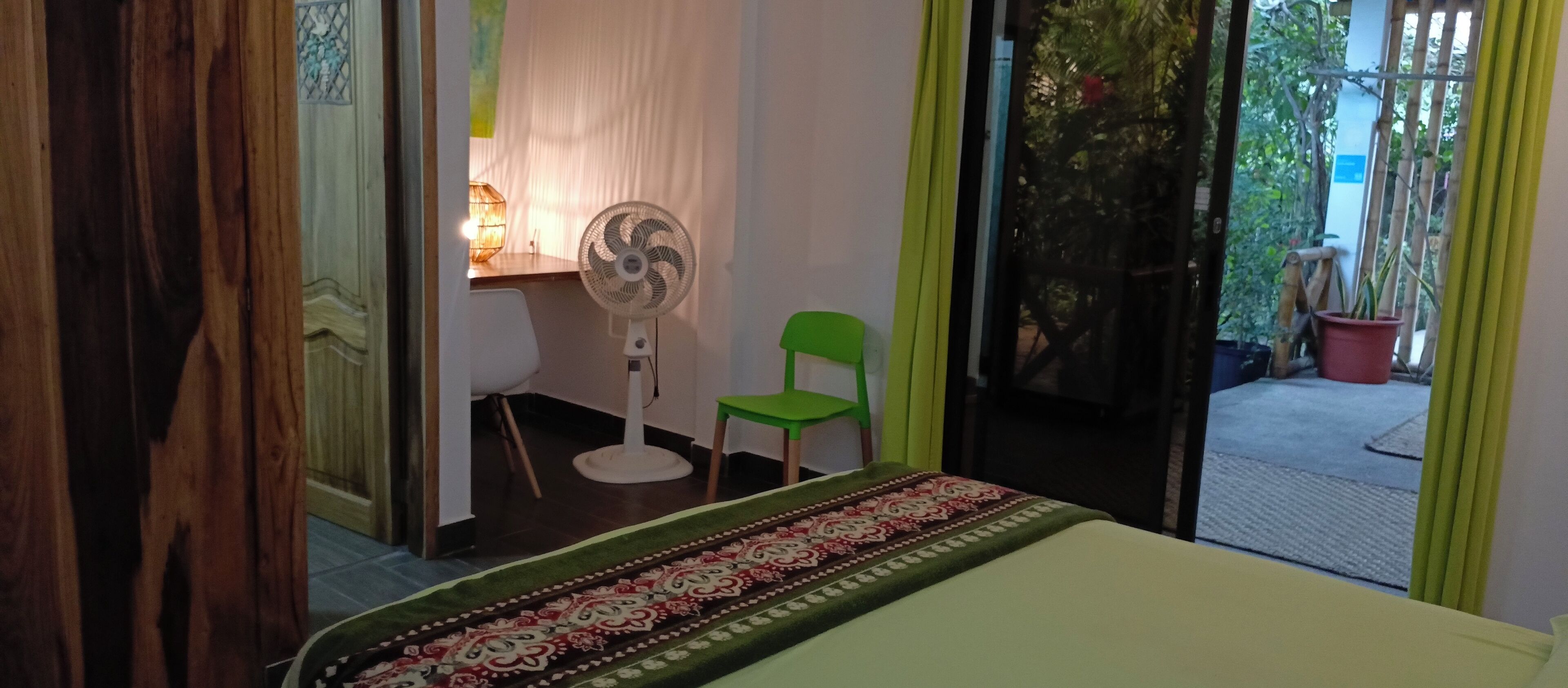 Una habitación con una cama, un ventilador, una silla verde y vistas al exterior por una puerta de cristal.