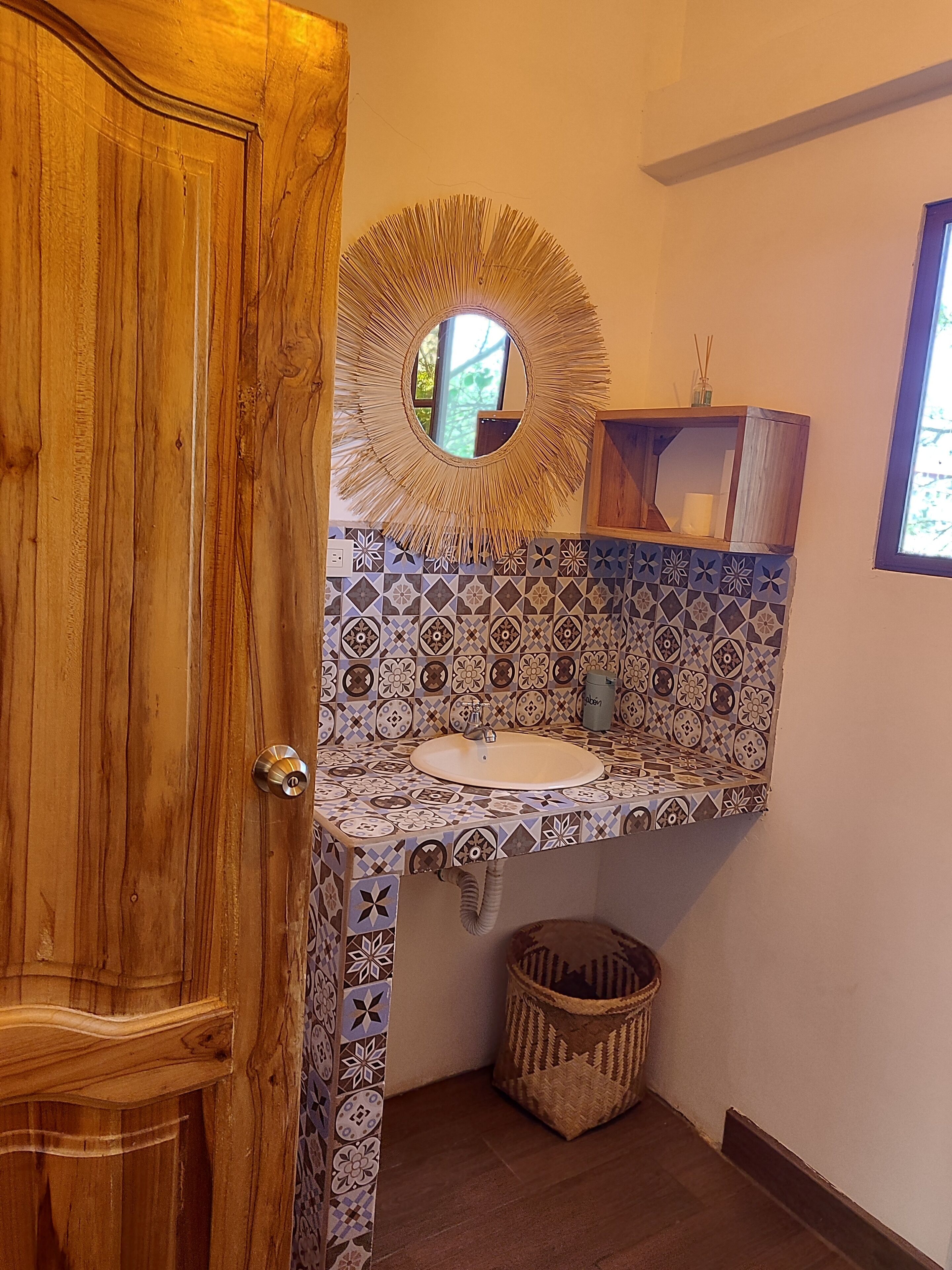 Une salle de bain avec une porte en bois, un lavabo agrémenté d’un miroir décoratif, des carreaux à motifs, une petite étagère et une poubelle.