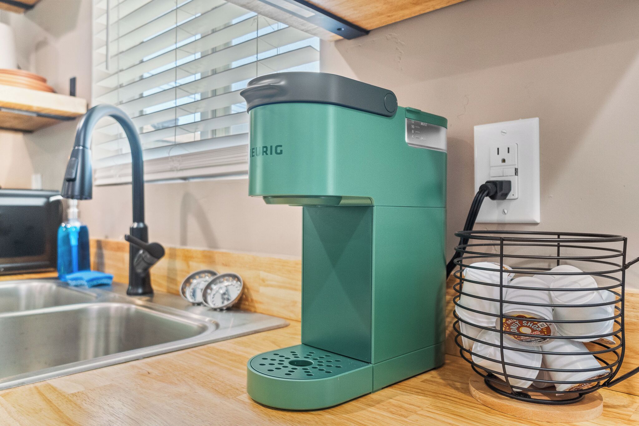 Una cafetera verde sobre una encimera de cocina con un fregadero, un reloj y un cesto de cápsulas de café.