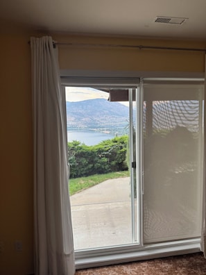 A room with a large sliding glass door leading to a balcony with a view of a lake and mountains.