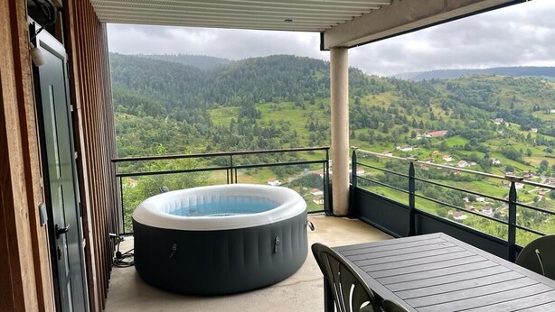 A balcony with a hot tub, a table, and a view of a green valley. - LE Massif - Appartement Avec vue Imprenable (La Bresse)