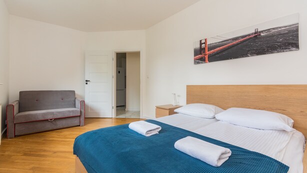 A hotel room with a bed, a grey sofa, a wooden nightstand, and a painting of a bridge. - Molo Centrum by Grand Apartments (Sopot)