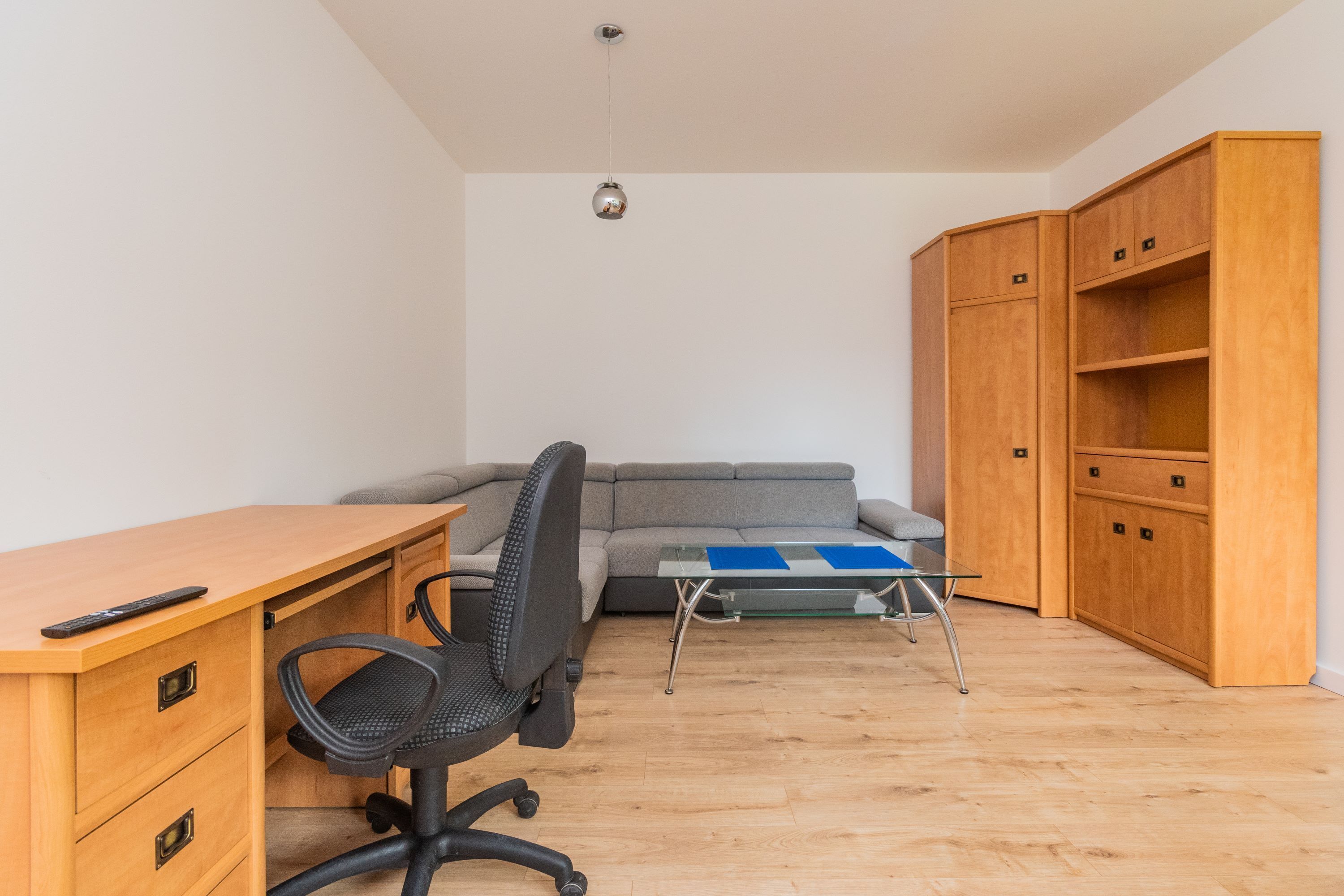 Una habitación compacta con un escritorio de madera, una silla de oficina negra, un sofá gris y una pequeña mesa de vidrio.