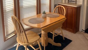 A dining area with a wooden table, four chairs, a blue rug, and a kitchen with wooden cabinets and a hanging light fixture. - The Hideaway Cabin #63 (Gold Beach)