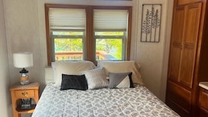 A bedroom with a large bed, two windows, a nightstand with a lamp, and a wooden wardrobe. - The Hideaway Cabin #63 (Gold Beach)
