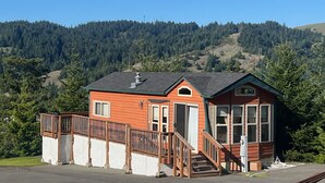 A small, single-story building with a red exterior and a wooden deck, surrounded by dense forest and hills. - The Hideaway Cabin #63 (Gold Beach)