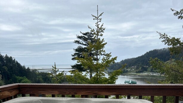 A hot tub on a deck with a view of a lake, trees, and a bridge in the background. - The Hideaway Cabin #63 (Gold Beach)