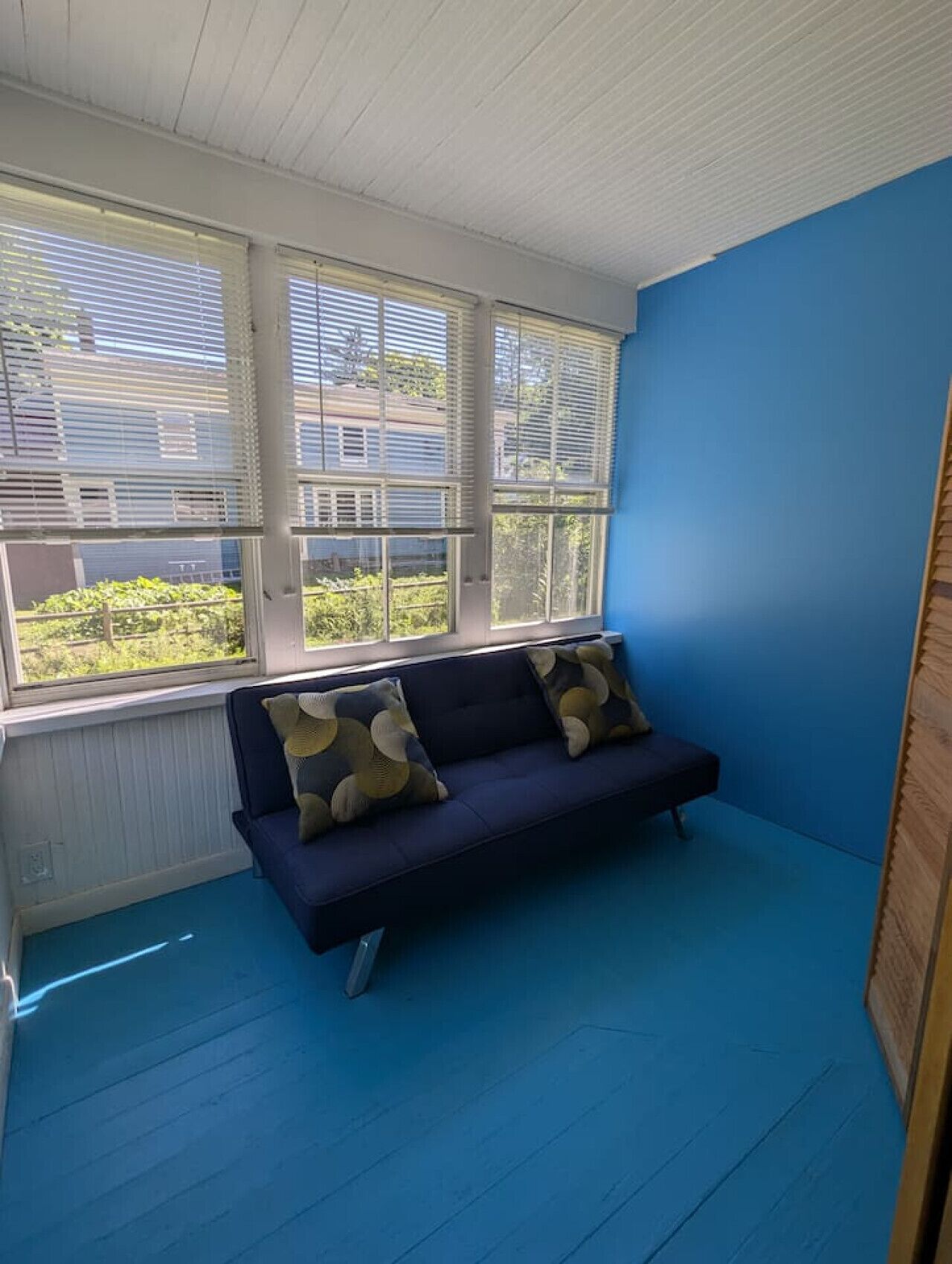 Une pièce avec un mur bleu, un canapé bleu foncé orné de coussins à motifs et une fenêtre avec des stores.