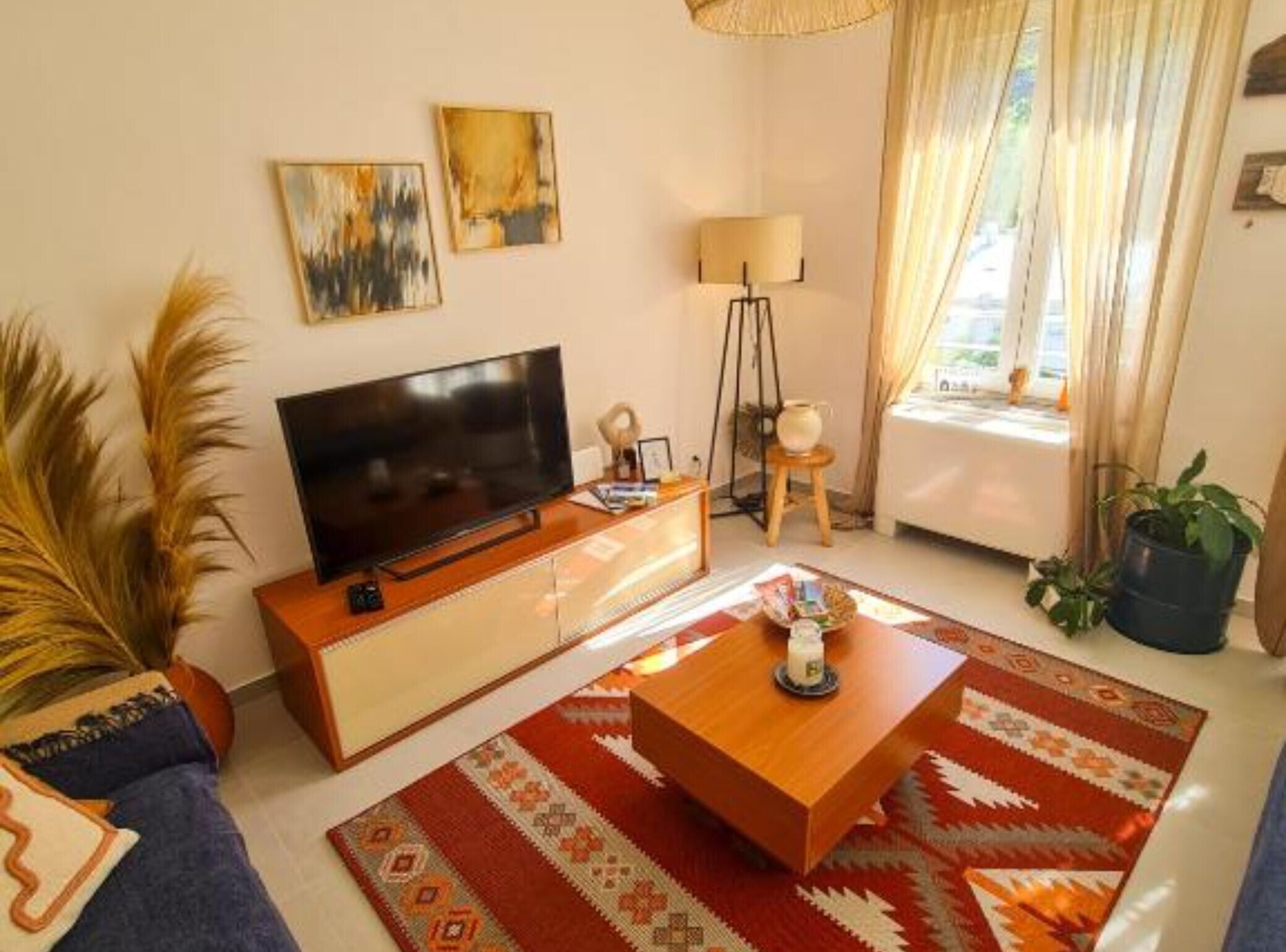 A living room with a flat-screen TV, a coffee table, and a sofa. There are two paintings on the wall, a lamp, and a window with curtains.