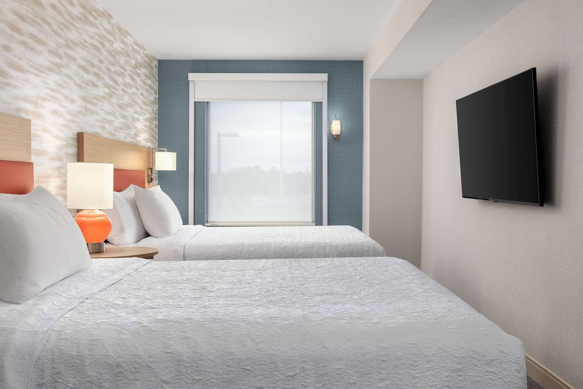 A hotel room with two beds, a large window with blinds, a flat-screen TV mounted on the wall, and a nightstand with a lamp.