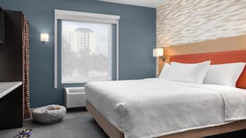 A hotel room with a bed, a pet bed, a window, a wall-mounted lamp, and a view of a building outside.