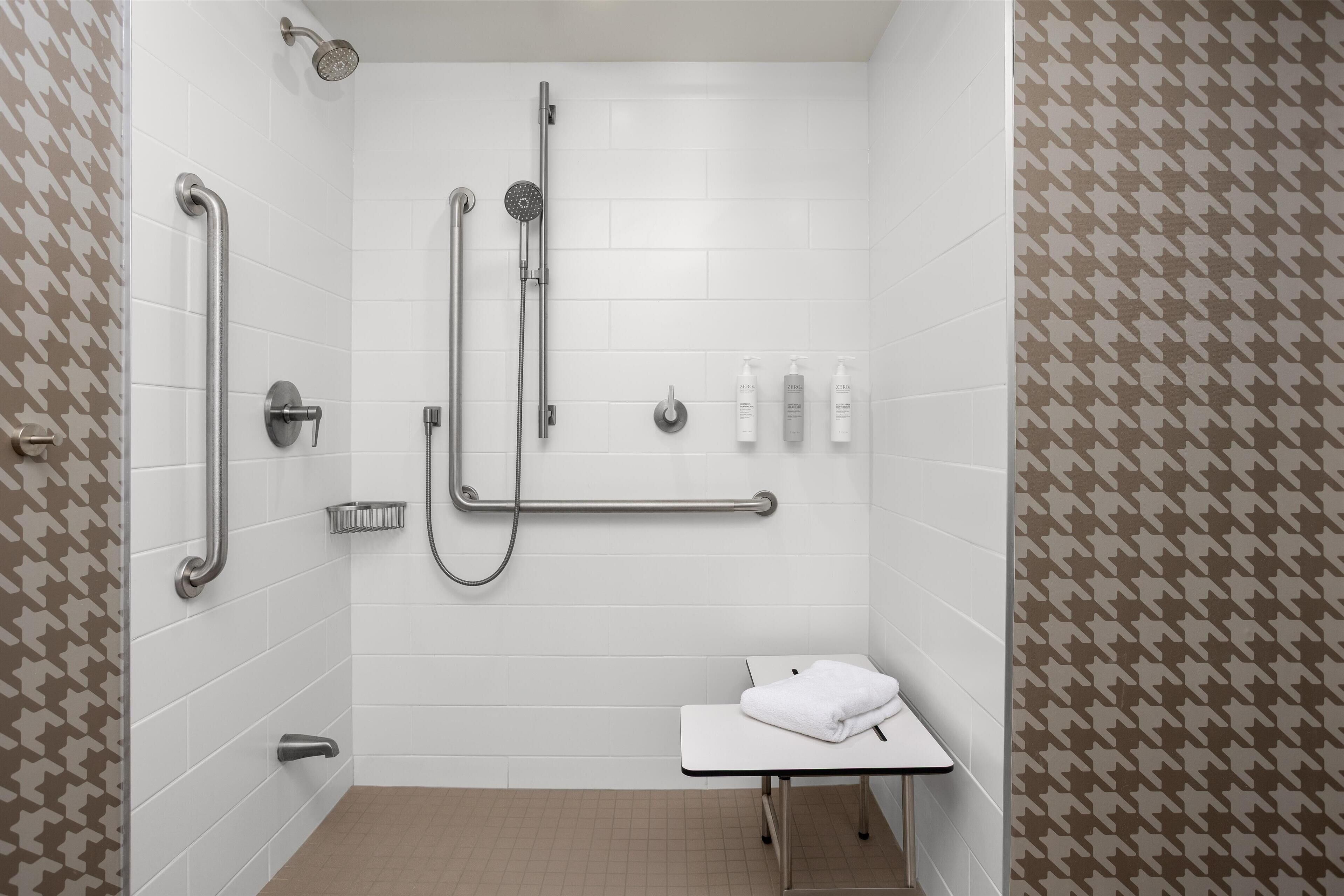 Un baño moderno con paredes de azulejos blancos, una ducha con cabezal de ducha manual, barras de apoyo y una mesita con una toalla.
